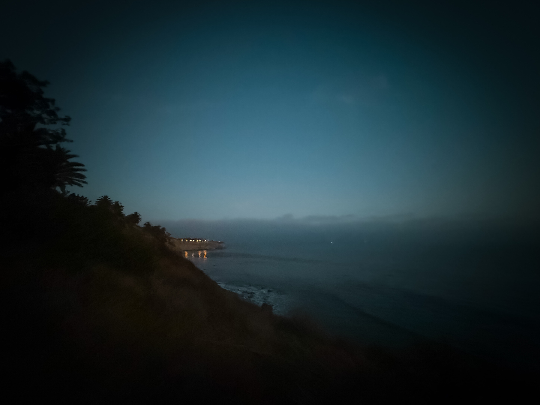 A serene coastal scene is captured at dusk, with faintly illuminated buildings in the distance and a calm ocean under a darkening sky.
