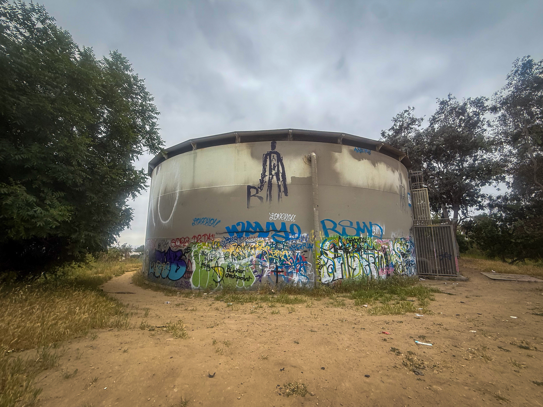 A large cylindrical structure covered in colorful graffiti is situated outdoors, surrounded by trees and grassy terrain.