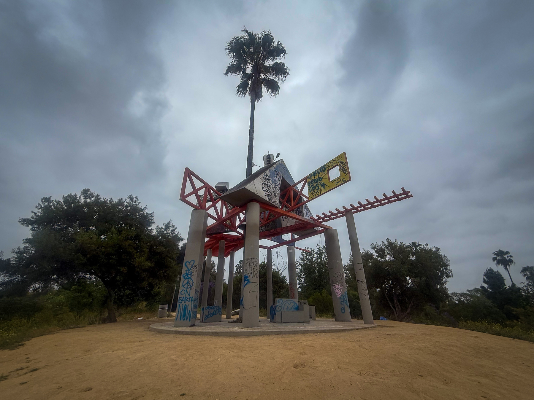 A unique, graffiti-covered structure with tall pillars and a central palm tree stands under a cloudy sky.