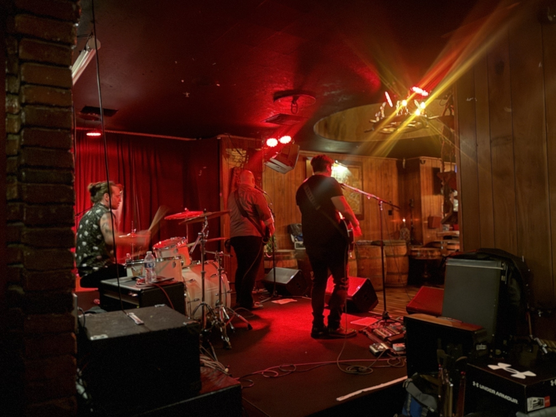 A band is performing on a dimly lit stage in a cozy venue with wooden walls and red lighting.