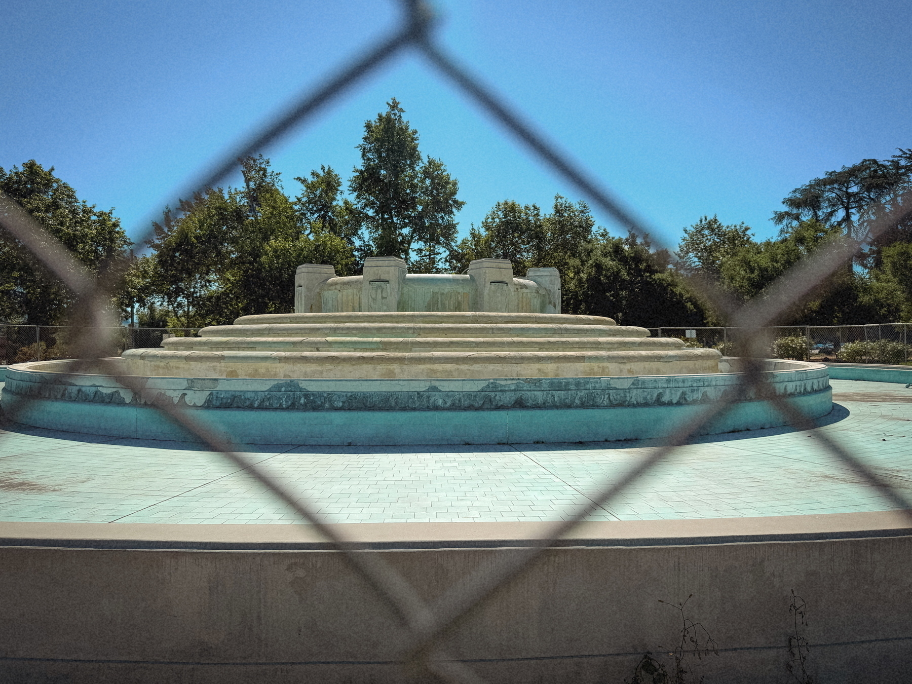 A dry, tiered fountain surrounded by trees is visible through a chain-link fence.