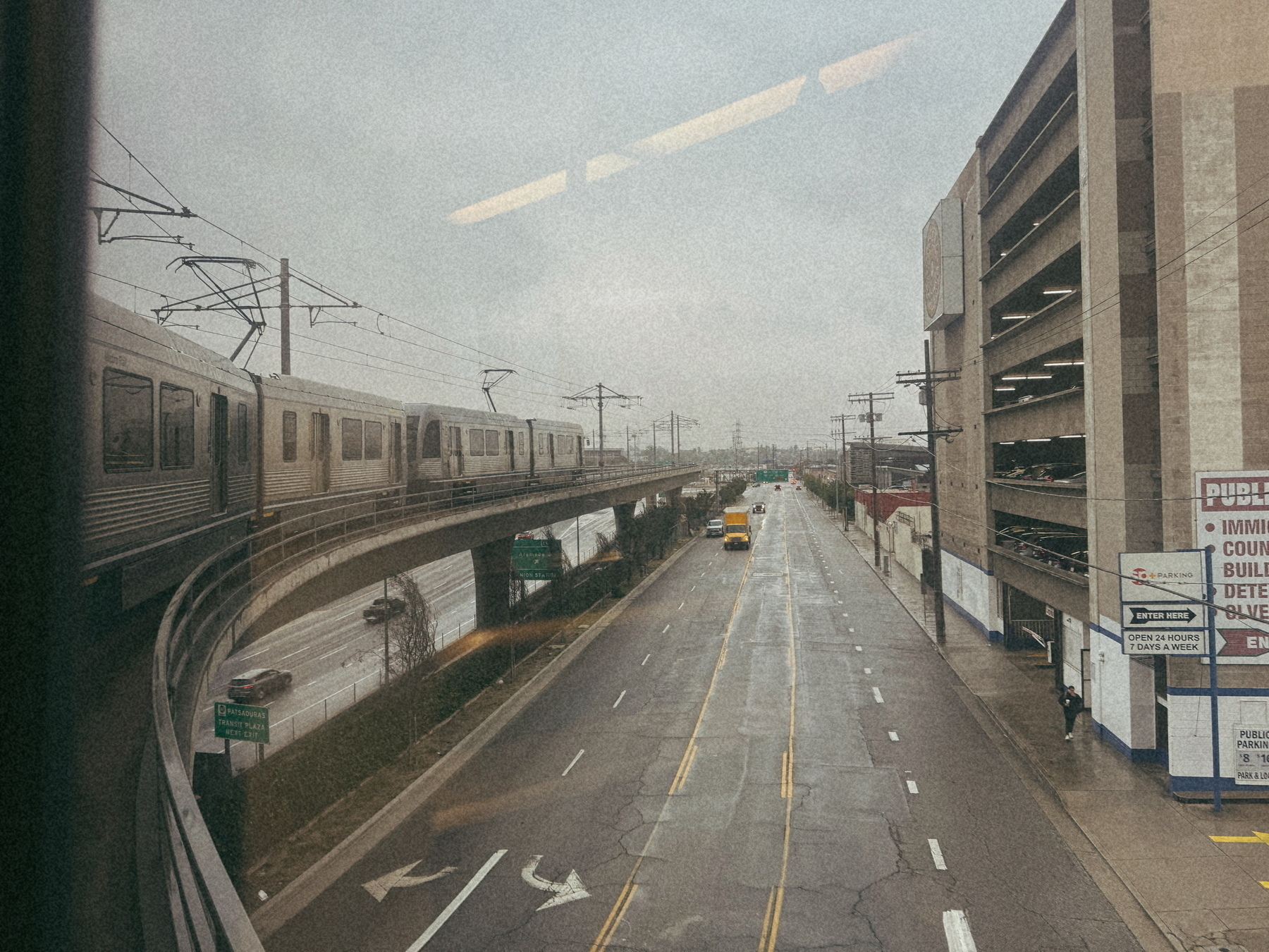 A train travels along an elevated track beside a quiet, overcast city street with a bus and a multi-story parking garage in the background.