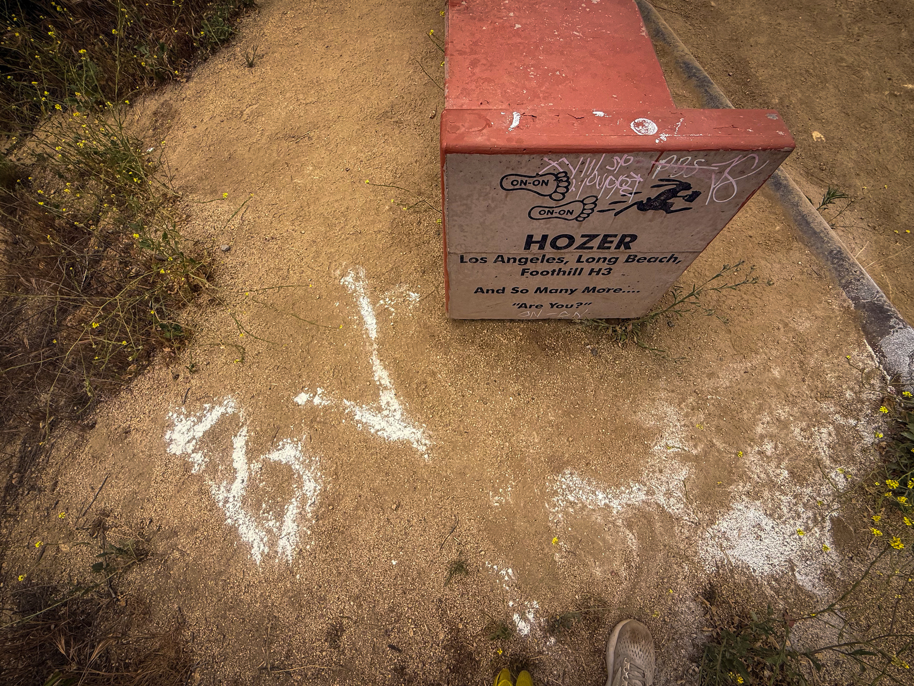 A trail marker is situated on a dirt path, displaying various drawings and writings, with some chalk markings on the ground nearby.